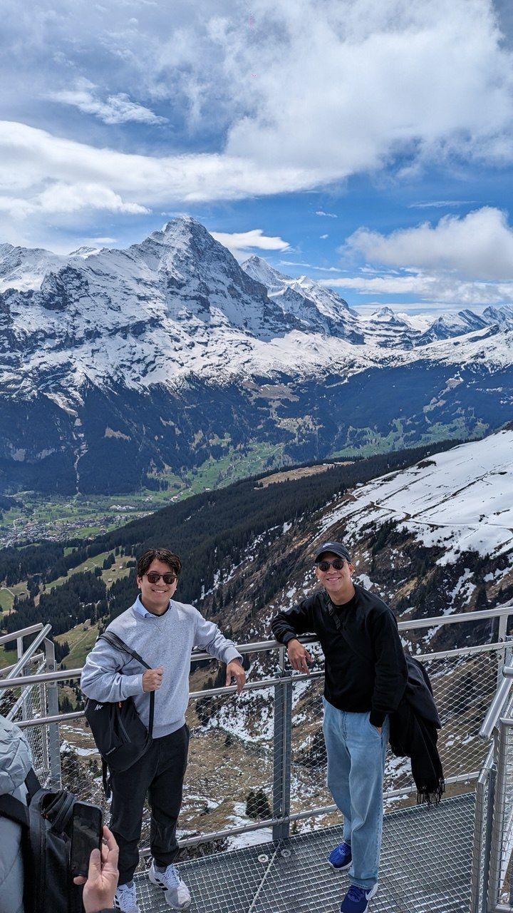 The twins smiling on a mountain viewing platform, with snowy peaks and a green valley behind them.